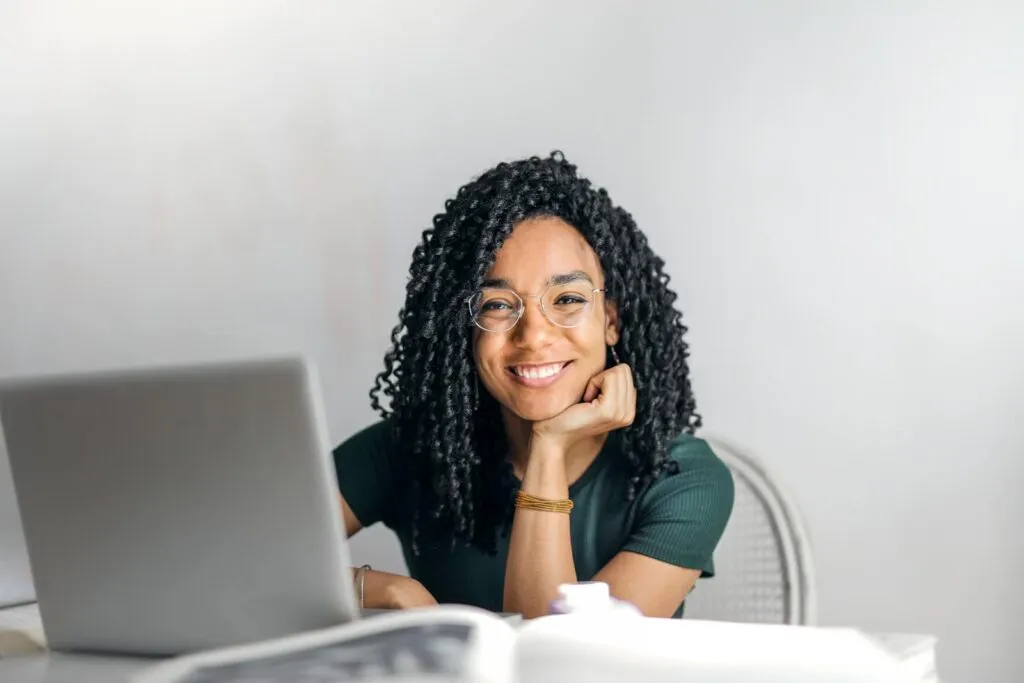 7 Vagas de Estágio Remoto e Híbrido Joyful businesswoman with curly hair smiling at camera while using laptop indoors.