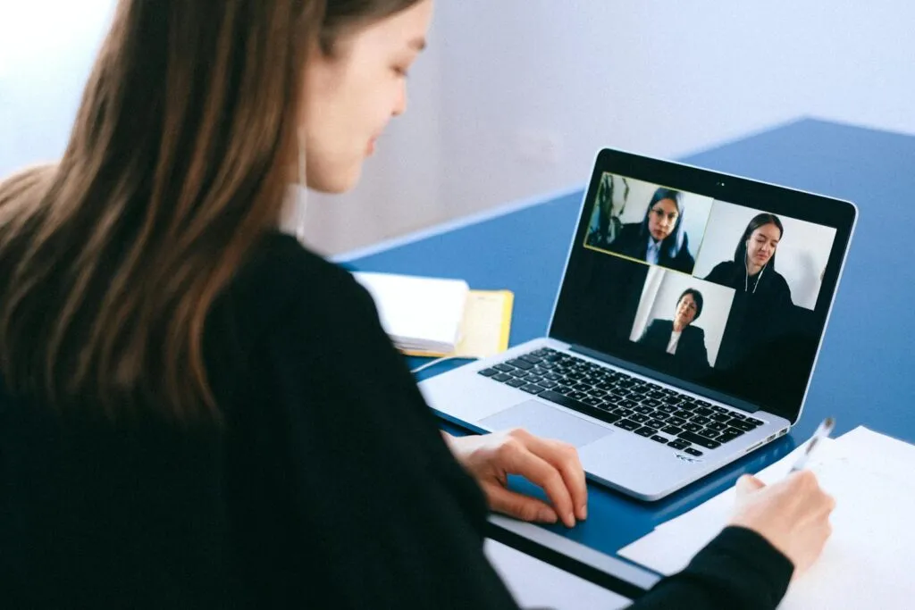 Vagas Remotas na Máquina de Vendas A woman engaging in a video conference using a laptop at home, taking notes.