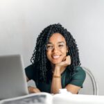 7 Vagas de Estágio Remoto e Híbrido Joyful businesswoman with curly hair smiling at camera while using laptop indoors.